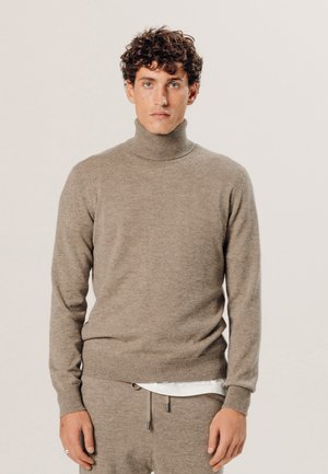 Beige Rollkragenpullover aus weichem Material, mit langen Ärmeln und gerippten Bündchen, kombiniert mit passenden Hosen und einem weißen Hemd darunter.