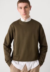 Sweat-shirt vert olive avec un col rond, arborant un logo de marque en relief. Superposé à une chemise blanche à col, sur un fond neutre.