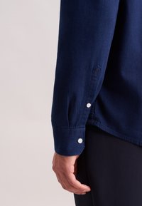 Mano maschile appoggiata di lato, che indossa una camicia a maniche lunghe blu navy con bottoni bianchi e pantaloni scuri su uno sfondo neutro.