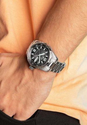 Montre en acier inoxydable argenté avec un cadran noir, de grands chiffres arabes et un bracelet en maillons métalliques. L'affichage de la date est positionné à 3 heures.