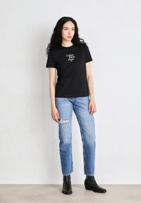 Juodas medvilnės marškinėliai su baltu "Calvin Klein Jeans NYC" grafiniu dizainu, derinami su šviesiai mėlynomis dėvėtomis džinsais ir juodais auliniais batais.