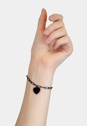 Bracelet en chaîne de métal noir avec un charme en forme de cœur au centre, doté de maillons lisses et d'une finition polie, porté sur un poignet de couleur claire.