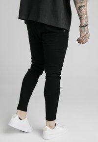 Jeans ajustados negros con una textura suave, que cuentan con un diseño de cinco bolsillos y costuras detalladas, combinados con zapatillas blancas.