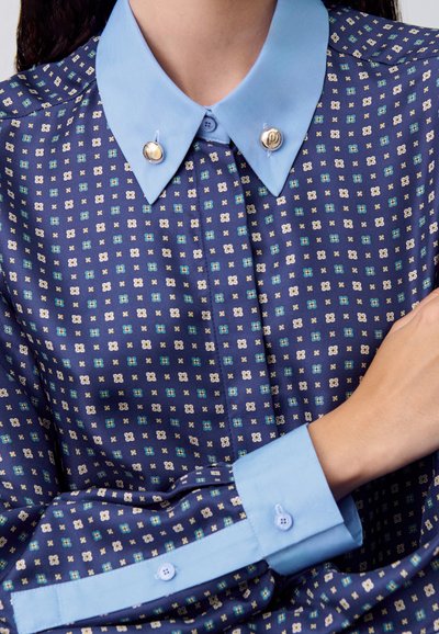 Camisa navy con un patrón geométrico en blanco, verde y azul. Presenta un color y puños en azul claro con botones decorativos.