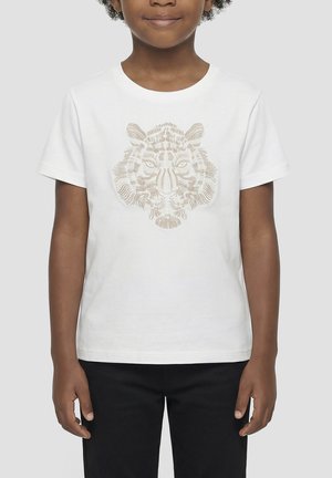 Enfant portant un t-shirt blanc avec un motif brodé beige de visage de tigre, associé à un pantalon noir, debout sur un fond uni.