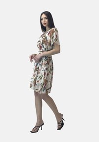 Robe fleurie à manches courtes, taille cintrée, tissu léger sur fond blanc, ornée de motifs de fleurs colorées. Talons noirs à lanières.