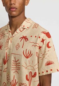Korte mouwen overhemd in beige stof met rode abstracte patronen, waaronder ontwerpen van de zon, cactus en palm. Met knoopsluiting en een gestructureerde weving.