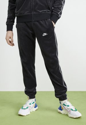 Person trägt schwarze Nike-Trainingshose mit weißen Seitenstreifen und weiße Sneaker mit grünen und blauen Akzenten, steht auf grünem Boden.