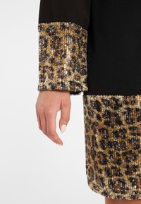Schwarzes Kleid mit goldfarbenem Leopardenmuster und Paillettenverzierungen an den Ärmeln und dem Saum. Glatter Stoff mit funkelnden Akzenten, die ein strukturiertes Aussehen erzeugen.