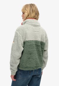 Fleece-pullover i lysegrå og grøn, med høj krave og ribbede ærmeafslutninger. Blød tekstur med en markant rød detalje på kraven.
