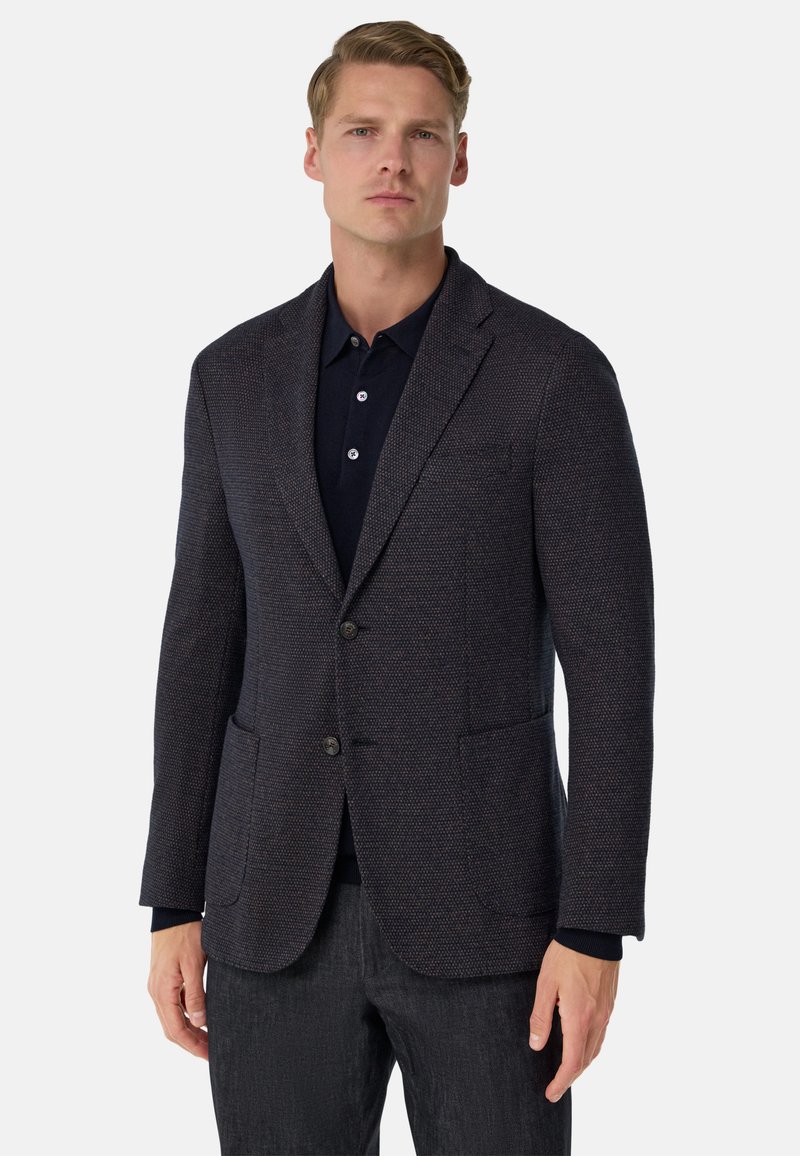 Blazer texturé bleu marine avec deux boutons, revers crantés et poches avant. Porte sur un polo sombre, associé à un pantalon foncé.