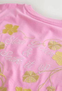 Roza bombažna majica z belim in zlatim vezenim napisom »Upočasni in zajemi trenutek« ter zlato cvetličnimi okraski.