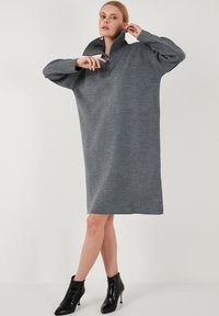 Grau oversized Kleid aus weichem Strickmaterial, mit hohem Kragen und einem Reißverschluss am Halsausschnitt. Kombiniert mit schwarzen Stiefeletten.