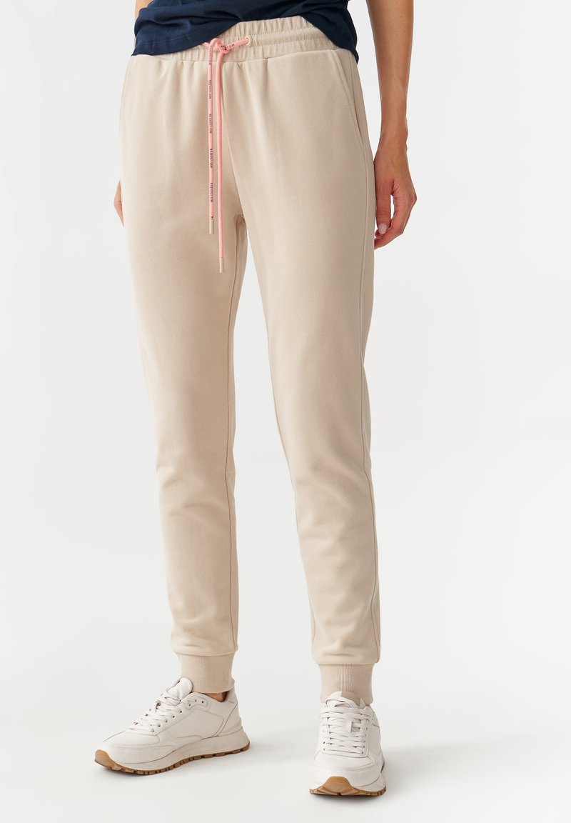 Beige joggerbyxor i ett mjukt material med dragsko i midjan, med en kontrasterande rosa snodd och ribbade muddar. Matchas med vita sneakers.