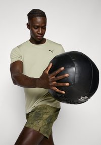 Puma kortermet oliven skjorte med svart logo, kombinert med mønstrete grønne shorts og en svart teksturert medisinball holdt i begge hender.