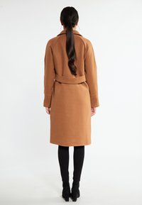 Cappotto marrone con cintura e finitura liscia e texture. Caratterizzato da un colletto e maniche lunghe, che raggiungono il polpaccio, abbinato a leggings neri e stivali con tacco.