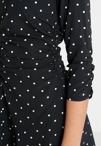 Černé šaty s bílými puntíky, se sebranými bočními detaily a nařasenými třičtvrtinovými rukávy, vyrobené z lehké látky.