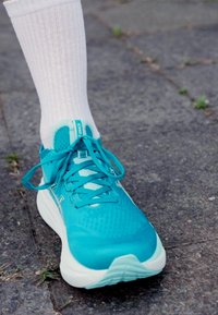 Turquoise athletic shoe with mesh upper, flat laces, and textured sole, worn with a white ribbed sock, on a stone pavement surface.