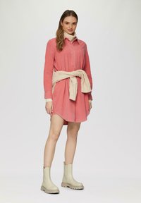 Abito camicia in corduroy rosa con una vestibilità rilassata, caratterizzato da una chiusura con bottoni sul davanti, maniche lunghe e un maglione beige in maglia legato in vita.
