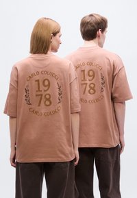 Πουκάμισο σε πολύ μεγάλο μέγεθος, ανοιχτό καφέ, από βαμβάκι. Διαθέτει εμπρόσθιο σχέδιο με την επιγραφή "CARLO COLUCCI 1978" με φυλλώδη διακοσμητικά σε πιο σκούρο καφέ.