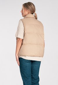 Beżowa pikowana kamizelka z wysokim kołnierzem, cechująca się gładką fakturą i bezrękawowym krojem. Nosi ją nadwym beige swetrem dzianinowym i niebieskimi spodniami o fakturze.