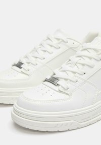 Weiße Sneakers mit glattem Lederobermaterial, gepolstertem Zungenbereich und Metallakzent an den Schnürsenkeln. Abgerundete Zehenkappe und strukturierte Gummisohle.