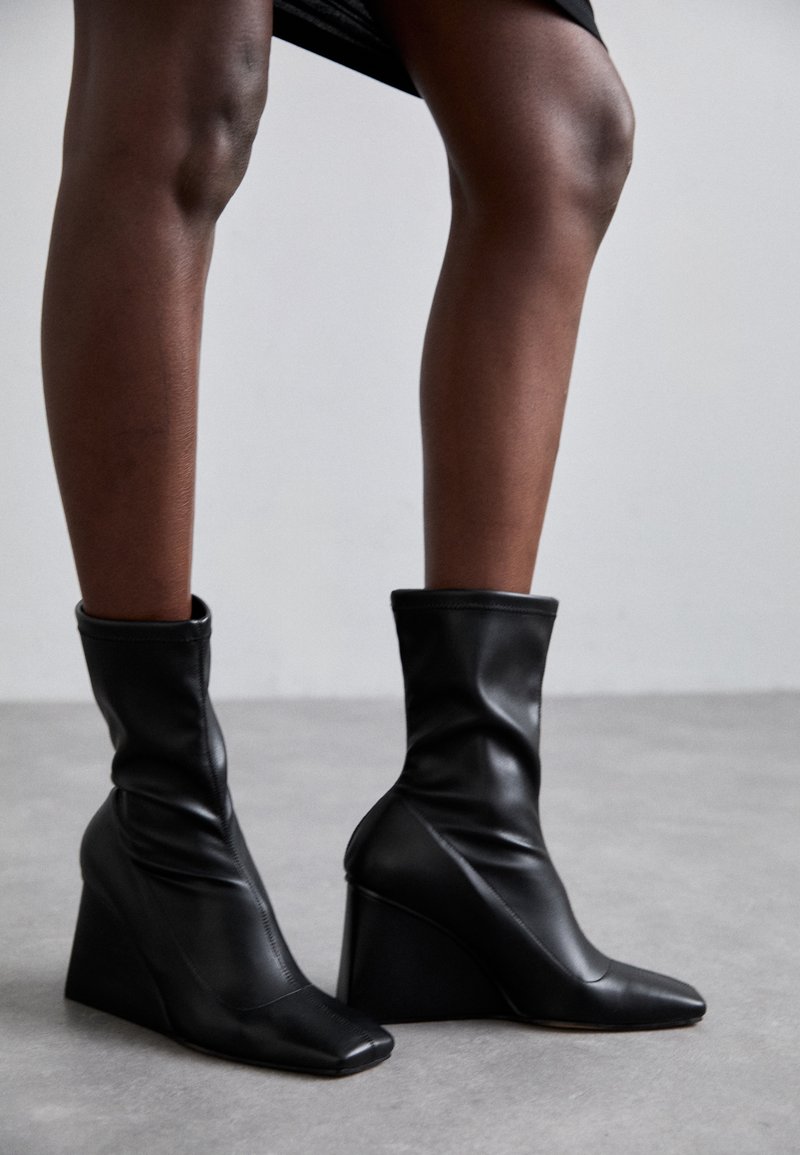 Cizme negre din piele cu toc wedge, cu un design lejer în partea superioară, vârf ascuțit și un toc unghiular distinctiv, evidențiind elemente de design modern.