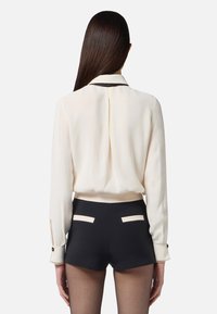 Blusa crema con colletto nero, maniche lunghe. Pantaloni corti neri con due accents chiari sulle tasche posteriori. Tessuto liscio, vestibilità su misura.