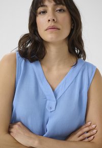 Blouse sans manches bleu clair avec un tissu texturé, un décolleté en V et un détail à bouton simple à l'avant.