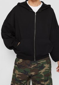 Felpa nera con zip e cappuccio, vestibilità oversize e tasche frontali; abbinata a pantaloni cargo con motivo camouflage.