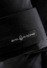 Zwarte stof close-up met een gestructureerde afwerking en een opvallend wit "SAIL RACING" logo op een rechthoekig label.
