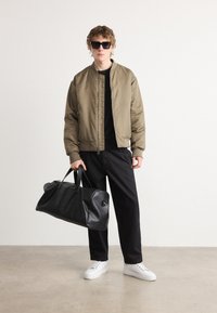 Veste bomber kaki, t-shirt noir, pantalons noirs, sneakers blancs, tenant un sac de voyage en cuir noir et portant des lunettes de soleil foncées.