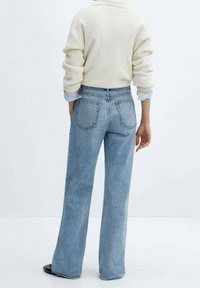 Persona vestida con jeans de pierna ancha en azul claro y un suéter de punto color crema, de pie de espaldas a la cámara sobre un fondo blanco liso.