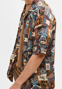 Chemise à manches courtes en tissu à motif géométrique multicolore bleu, marron et crème, avec col cubain et boutons.