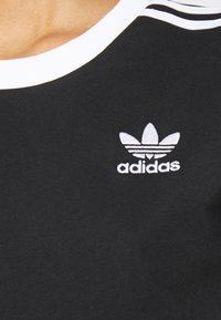T-shirt preto com um gola redonda branca e três riscas brancas no ombro, com um logo Adidas branco bordado no peito.