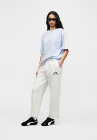 Ανοιχτό μπλε φαρδιά μπλούζα με λευκά μανίκια, συνδυασμένη με γκρι φόρμα με μαύρο λογότυπο "NEW BALANCE", φορεμένη με μαύρα αθλητικά παπούτσια.