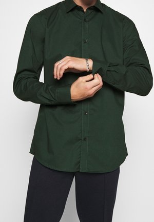 Mann trägt dunkelgrünes Hemd mit Knöpfen und schwarze Hose, richtet Hemdmanschette, trägt silbernes Kettenarmband und Ring an der linken Hand.