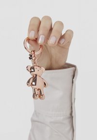 Llavero de metal color oro rosa con un acabado suave, unido a un llavero circular. Mano sosteniendo el llavero.