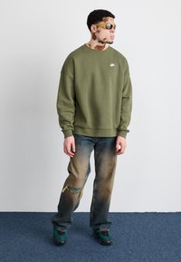 Green sweatshirt with white logo, distressed blue jeans, and teal sneakers. Accessories include yellow sunglasses. Plain background.