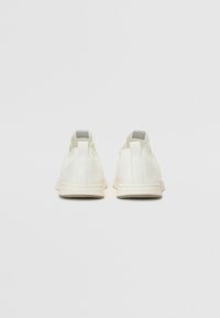 White sneakers with a smooth texture, minimalistic design, rounded shape, and a light-colored sole; includes a gray tab at the heel.