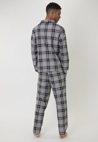 Man in een grijs en zwart geruite lange mouwen top en bijpassende broek staat blootvoets met handen in de zakken, met de rug naar de camera gekeerd tegen een eenvoudige achtergrond.