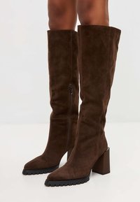 Stivali alti in suede marrone, con punta a punta, tacco a blocco e zip laterale. Superficie testurizzata e suola in gomma con motivo a battistrada.