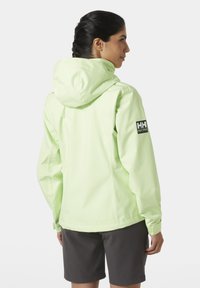 Chaqueta impermeable verde claro con capucha, con puños elásticos y un parche con el logo en la manga izquierda. Lleva puestos pantalones cortos oscuros.