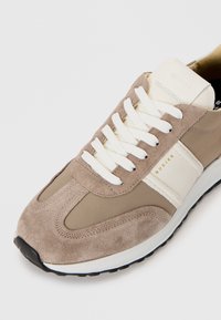 Beige sportsko med sneaks som har overdel i semsket skinn og tekstil, hvite snørebånd, og en teksturert svart såle med merkevare på siden og tungen.