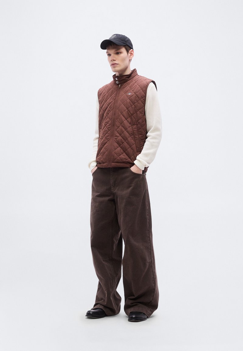 Quilted brown vest over a cream textured sweater, paired with loose brown pants and a black cap. Simple and functional design, no patterns.
