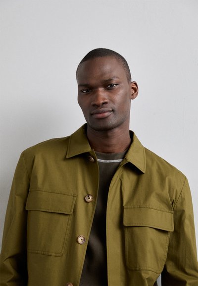 Olivengrøn jakke med knaplukning foran, to brystlommer og klassisk krave. Båret over en ribbet grå skjorte. Glat tekstur.