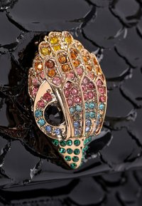 Spilla dorata a forma di mano, adornata con cristalli multicolori, con accenti di pietra nera e posizionata su una superficie nera testurizzata.