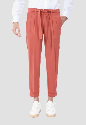 Persona che indossa pantaloni vita alta color ruggine con cintura annodata, camicia bianca abbottonata e sneakers bianche, in piedi su uno sfondo semplice.