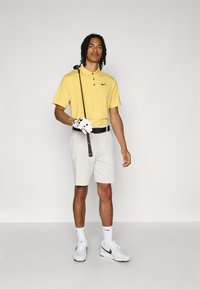 Πόλο μπλουζάκι σε κίτρινο χρώμα με τρία κουμπιά, συνδυασμένο με ανοιχτό μπεζ σορτς. Τα αξεσουάρ περιλαμβάνουν μια μαύρη ζώνη, ένα λευκό γάντι γκολφ και γκρίζα αθλητικά παπούτσια.