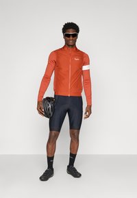 Costum de ciclism pentru bărbați, compus dintr-o jachetă de vânt roșie, cu croială slim-fit și accente albe, pantaloni scurți negri cu umplutură și pantofi de ciclism negri. Ține în mână un casca de bicicletă.